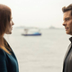 Christina and Teddy stare at each other on a pier while a boat passes between them in the distance.